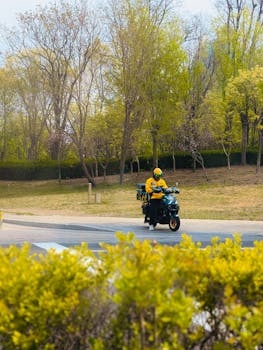 A courier in yellow uniform rides a scooter through a sunny park.