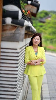 Asian woman in bright yellow suit stands confidently by an outdoor traditional wall.