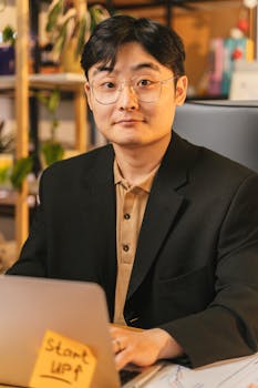Confident Asian businessman in eyeglasses working on a laptop with startup note, indoors.