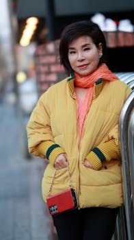 Asian woman in yellow jacket and orange scarf standing outdoors with hands in pockets.